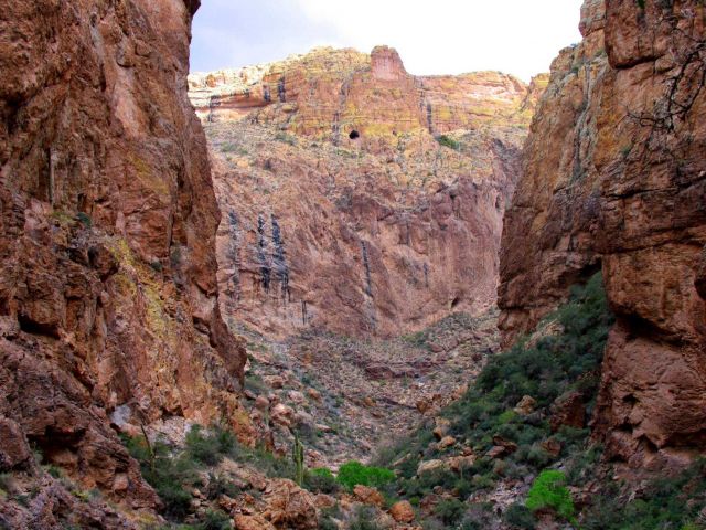 Sandstone cliffs, perhaps a lost mine on the cliff high above, and desert foliage in the canyon bottoms. Picture