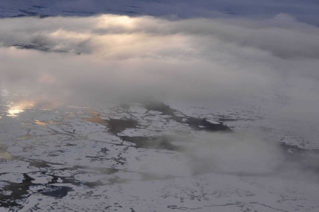 A helicopter eye's view of melting ice with clouds overhead illuminated through a layer of higher yet clouds. Picture