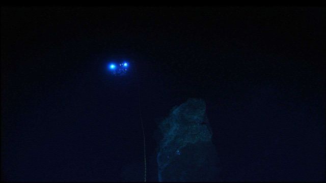 The lights from the Hercules ROV are seen 30-meters away as the ROV is maneueved in the vicinity of a rock outcrop. Picture