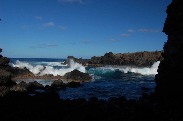 A small hole-in-the wall affords a glimpse of azure surf pounding against against jagged volcanic rocks. Picture