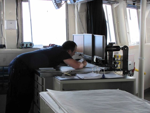 Although electronic charts are used, the officer of the deck is maintaining a separate paper chart plot of navigation. Picture