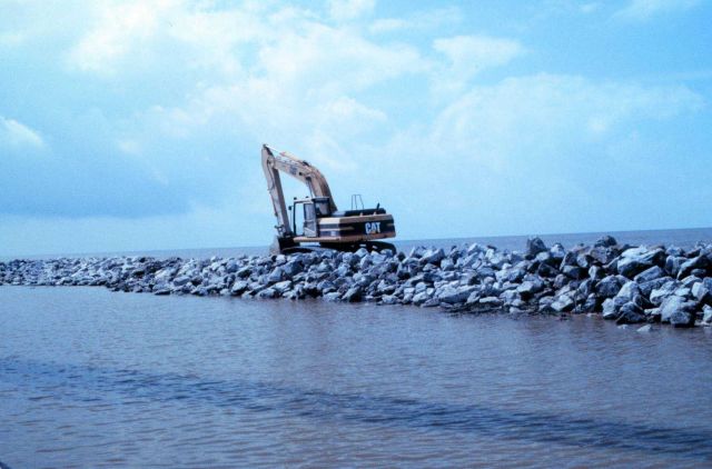 A view from the Mobile Canal of a back hoe placing rock at the narrow beach. Picture