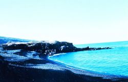 An arcuate cobble beach with a small volcanic rock outcrop at the end of the beach. Image