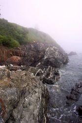 Quartz-veined metamorphic rocks meet the sea below a fog-shrouded lighthouse at West Quoddy Head. Image