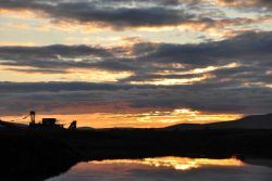 Sunset and clouds reflecting off water accented by the silhouette of an ancient gold dredge. Image